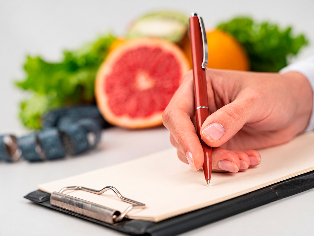 Profissional escrevendo em uma ficha de acompanhamento ao lado de alimentos frescos e um glicosímetro.