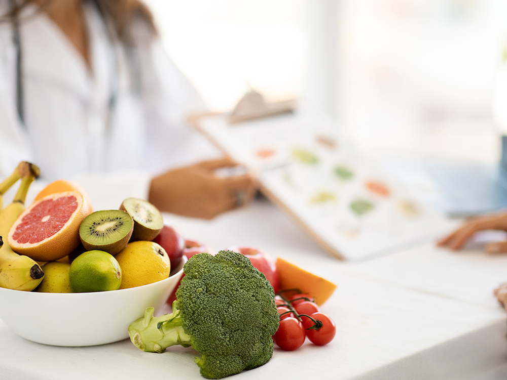 Nutricionista com prancheta ao fundo e, em primeiro plano, frutas, vegetais e tomates sobre a mesa de atendimento.