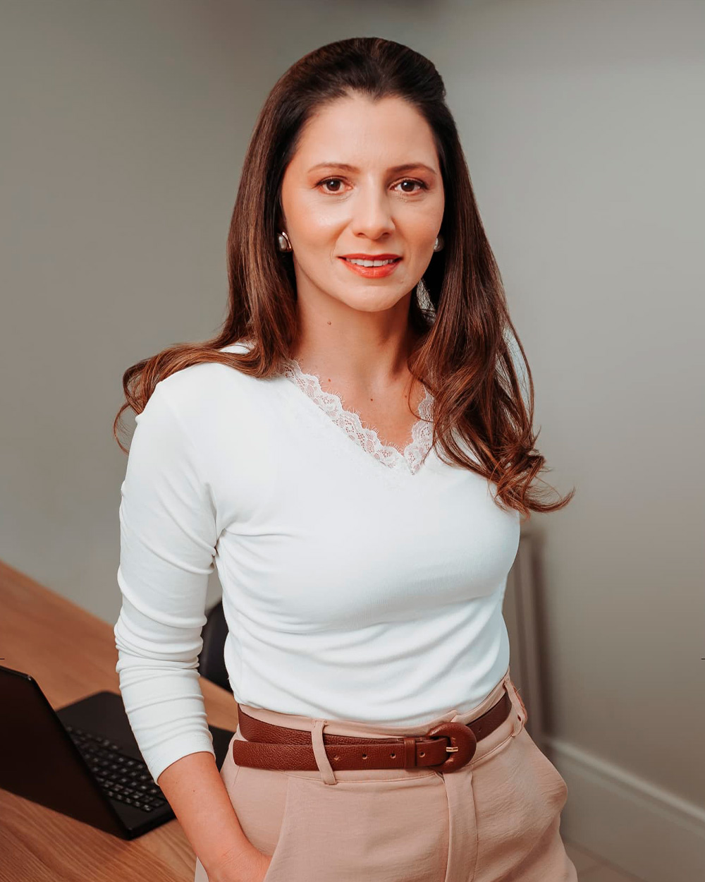 Fabiana Zanuzzo em pé no consultório, de blusa branca, junto à mesa de atendimento.